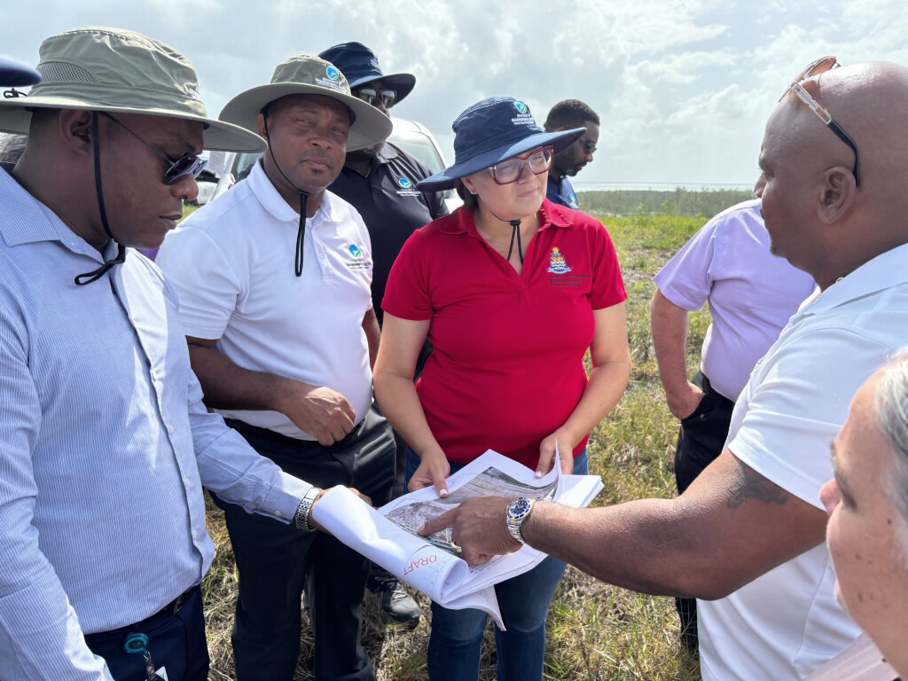 Minister Katherine Ebanks-Wilks wearing a red t-shirt and blue sun hat stands at the top of the George Town Landfil site in Grand Cayman with Department of Environmental Health Team members, looking at a report.